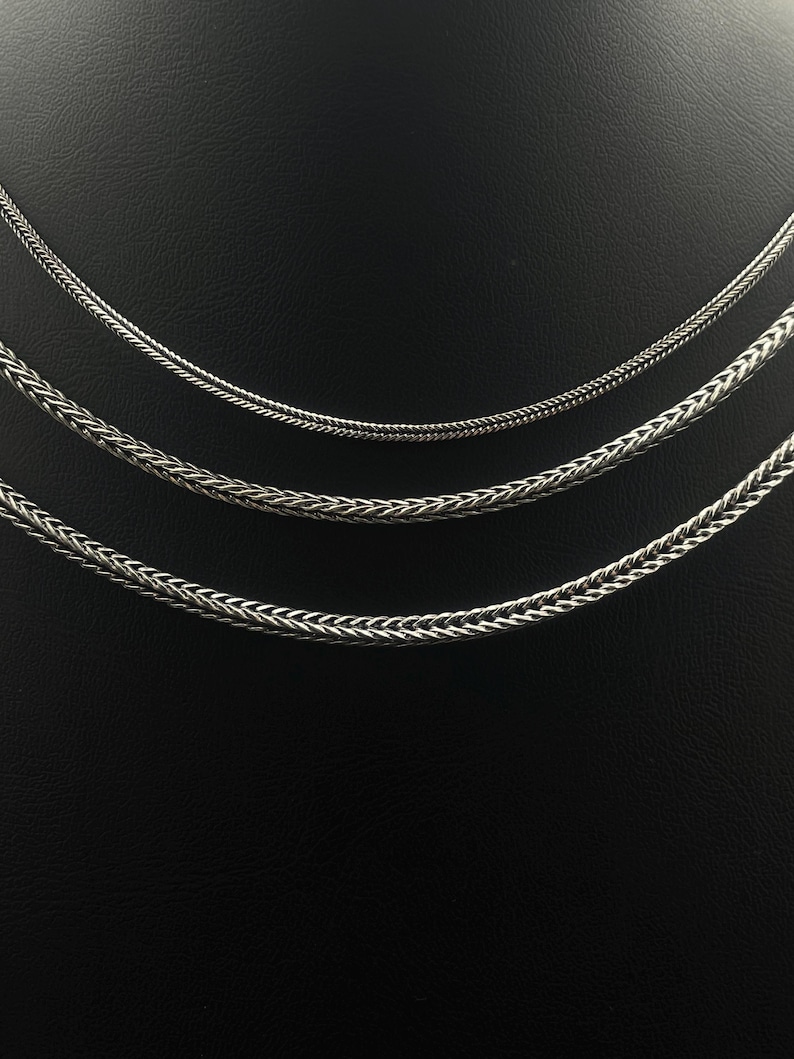 May include: Three silver-colored chain necklaces of varying thicknesses are displayed against a black background. The chains have different woven patterns, with the thickest chain at the bottom and the thinnest at the top. The chains are likely made of metal.