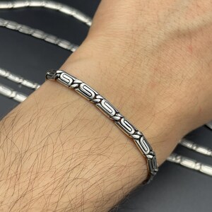 Puede incluir: Una pulsera de plata con un intrincado patr&oacute;n repetitivo de formas de 'S' estilizadas. La pulsera se lleva en una mu&ntilde;eca, mostrando su dise&ntilde;o y artesan&iacute;a. Joyer&iacute;a de plata.