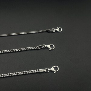 May include: Three silver-colored chain necklaces with lobster claw clasps are displayed against a dark background. The chains have different woven patterns, with one being a thinner, more delicate design.