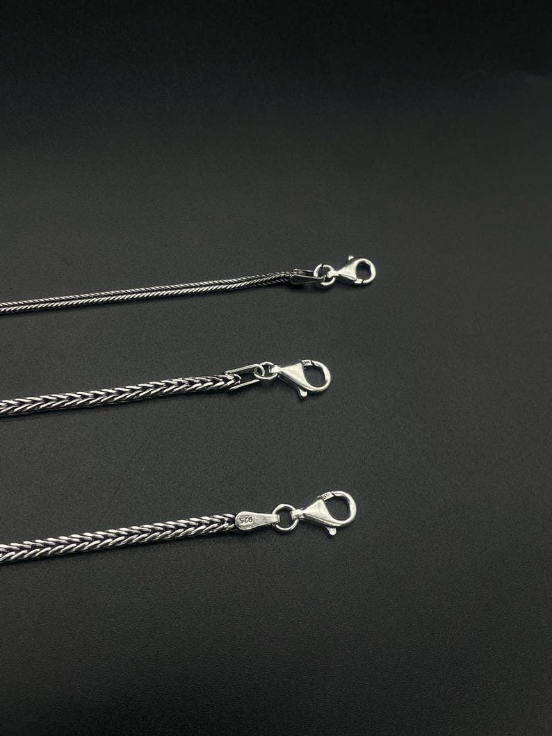 May include: Three silver-colored chain necklaces with lobster claw clasps are displayed against a dark background. The chains have different woven patterns, with one being a thinner, more delicate design.