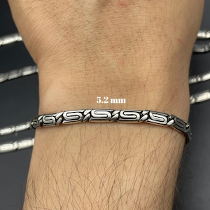 Puede incluir: Pulsera plateada con un dise&ntilde;o geom&eacute;trico repetitivo. La pulsera mide 5,2 mm de ancho y se muestra en una mu&ntilde;eca. La pulsera es de metal y tiene un acabado pulido. La pulsera es una joya.