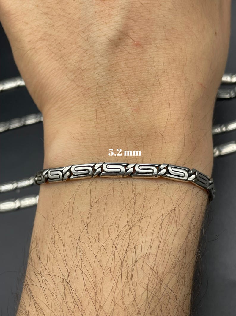 Puede incluir: Pulsera plateada con un dise&ntilde;o geom&eacute;trico repetitivo. La pulsera mide 5,2 mm de ancho y se muestra en una mu&ntilde;eca. La pulsera es de metal y tiene un acabado pulido. La pulsera es una joya.