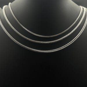 May include: Three silver-colored necklaces with different chain styles are displayed on a black mannequin. The necklaces are arranged in a layered fashion, showcasing varying textures and designs. The chains appear to be made of metal.