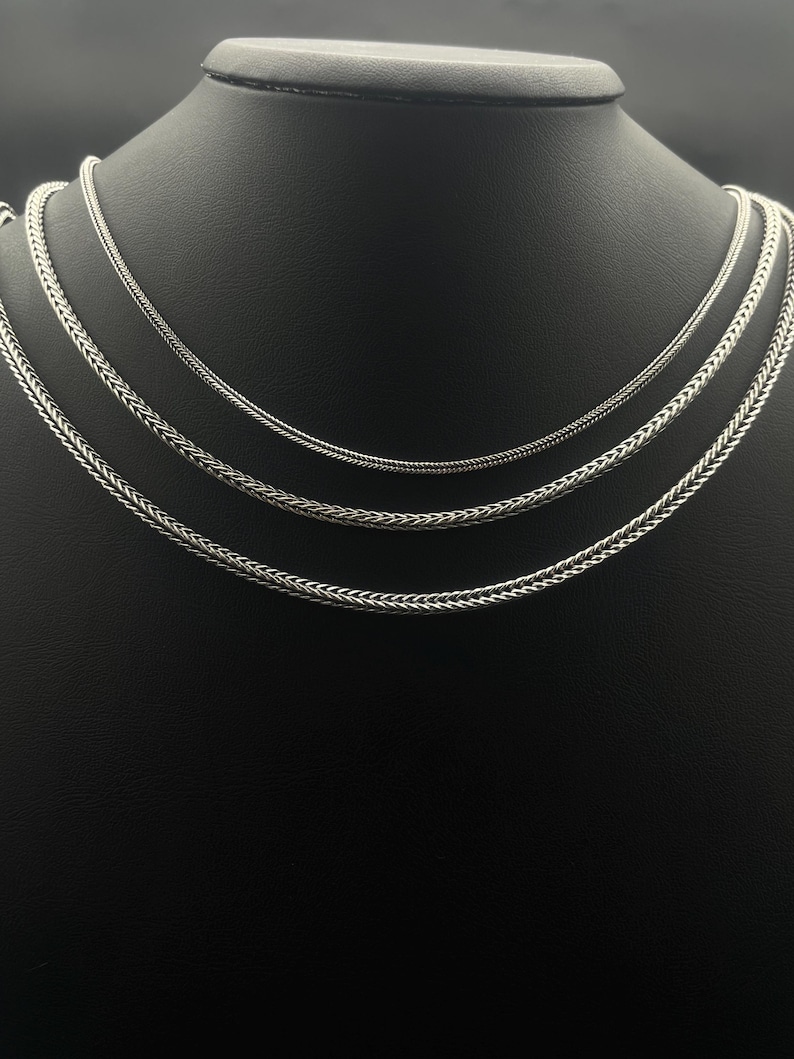 May include: Three silver-colored necklaces with different chain styles are displayed on a black mannequin. The necklaces are arranged in a layered fashion, showcasing varying textures and designs. The chains appear to be made of metal.