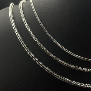 May include: Three silver-colored chain necklaces with a woven design are displayed on a black mannequin. The chains vary in thickness, with the thickest chain on the bottom and the thinnest at the top. The chains are made of a shiny metal.