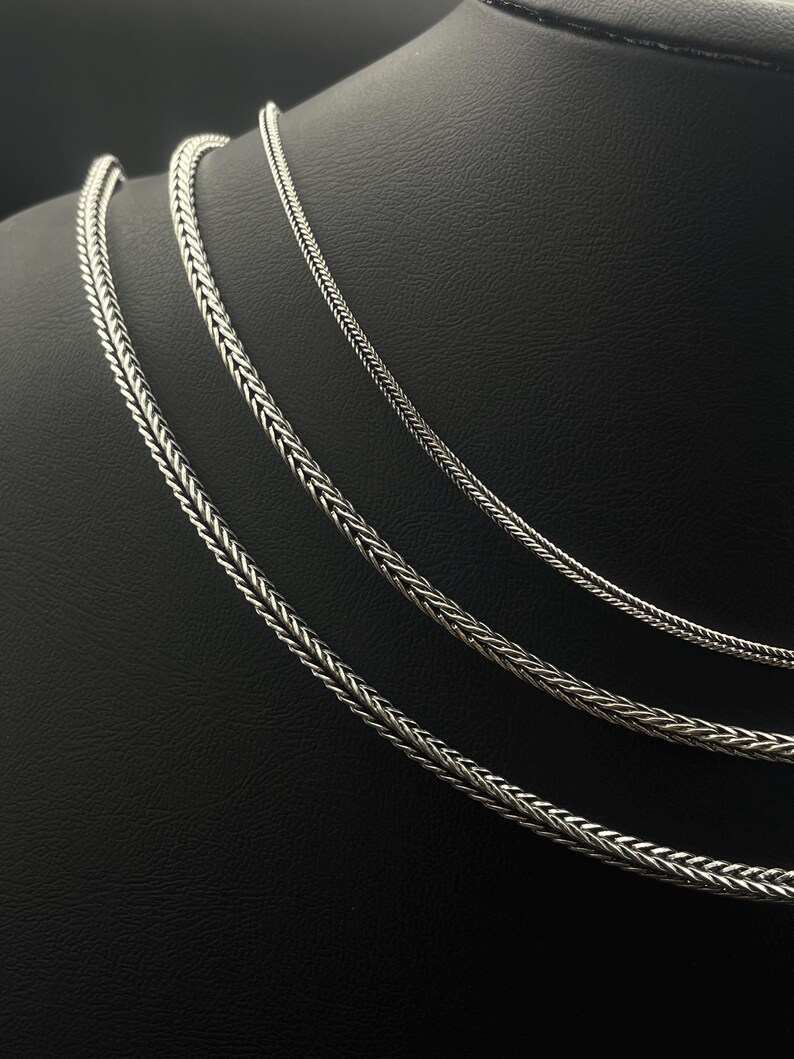 May include: Three silver-colored chain necklaces with a woven design are displayed on a black mannequin. The chains vary in thickness, with the thickest chain on the bottom and the thinnest at the top. The chains are made of a shiny metal.
