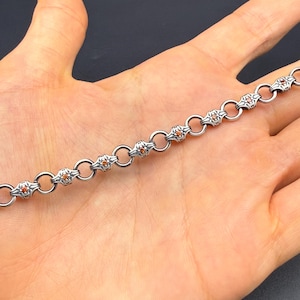 Könnte beinhalten: Ein silberfarbenes Metallkettenarmband mit sich wiederholenden dekorativen Gliedern in oxidierter Optik, die sich mit schlichten, runden Verbindungsringen abwechseln. Jedes dekorative Glied ist mit einem kleinen, runden, orangefarbenen Edelstein besetzt.
