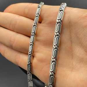Puede incluir: Un collar de cadena plateada con un dise&ntilde;o de eslabones en forma de S repetidos. El collar se muestra sobre un fondo oscuro. La cadena est&aacute; hecha de un material met&aacute;lico brillante.