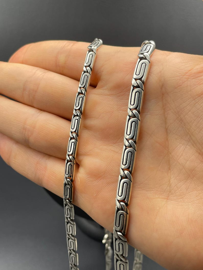 Puede incluir: Un collar de cadena plateada con un dise&ntilde;o de eslabones en forma de S repetidos. El collar se muestra sobre un fondo oscuro. La cadena est&aacute; hecha de un material met&aacute;lico brillante.