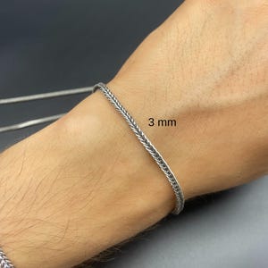 May include: A silver-colored chain bracelet with a woven design. The bracelet is 3 mm wide and is displayed on a person's wrist. The background is a dark gray color.