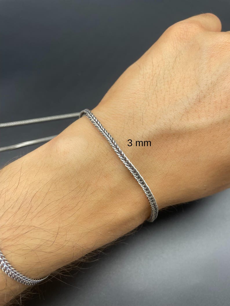 May include: A silver-colored chain bracelet with a woven design. The bracelet is 3 mm wide and is displayed on a person's wrist. The background is a dark gray color.