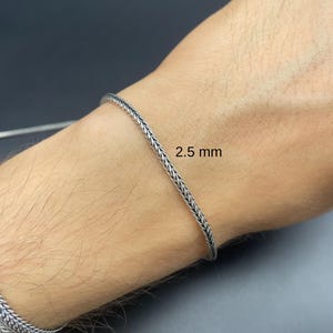 May include: A silver-colored bracelet with a braided design, 2.5 mm wide, is shown on a person's wrist. The bracelet has a textured appearance and is made of a shiny metal. The background is a dark gray color.