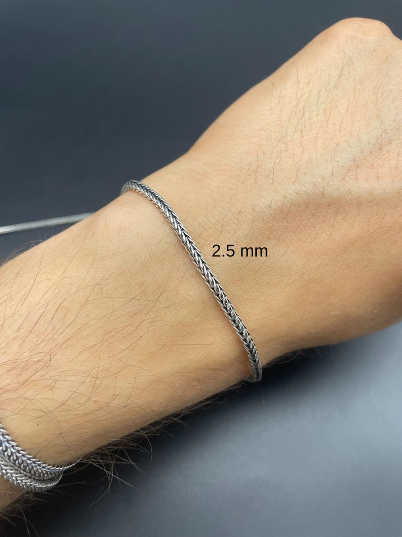 May include: A silver-colored bracelet with a braided design, 2.5 mm wide, is shown on a person's wrist. The bracelet has a textured appearance and is made of a shiny metal. The background is a dark gray color.