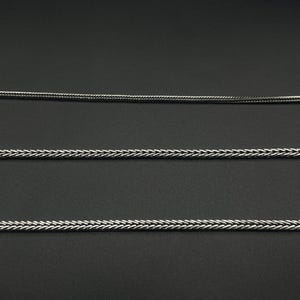 May include: Three silver-colored chain necklaces with a braided design are displayed against a dark background. The chains are of varying thicknesses, with a detailed weave pattern. The image highlights the texture and metallic sheen of the jewelry.