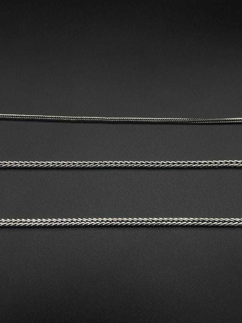 May include: Three silver-colored chain necklaces with a braided design are displayed against a dark background. The chains are of varying thicknesses, with a detailed weave pattern. The image highlights the texture and metallic sheen of the jewelry.