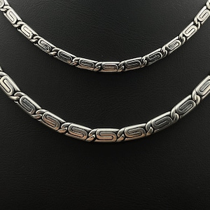 Puede incluir: Dos collares plateados con un dise&ntilde;o geom&eacute;trico repetitivo. Los collares se muestran sobre un fondo negro. Los eslabones tienen forma rectangular con un dise&ntilde;o estilizado de 'S' en el centro.