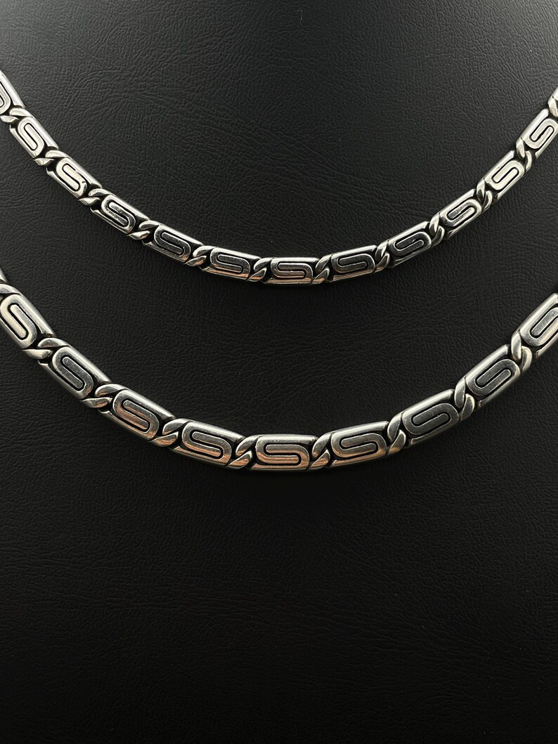 Puede incluir: Dos collares plateados con un dise&ntilde;o geom&eacute;trico repetitivo. Los collares se muestran sobre un fondo negro. Los eslabones tienen forma rectangular con un dise&ntilde;o estilizado de 'S' en el centro.