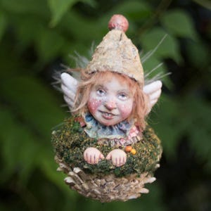 May include: A charming fairy ornament with a smiling face, freckles, and small white wings. The fairy wears a textured hat and a mossy green dress. The ornament is set against a blurred green background.