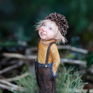 Puede incluir: Una figura caprichosa de una pequeña criatura con un sombrero en forma de piña, que lleva una camisa de color mostaza, un mono marrón y zapatos negros. La figura tiene una expresión sonriente y está de pie en una zona cubierta de hierba. El nombre del artista, TATJANA RAUM, está en la parte superior de la imagen.