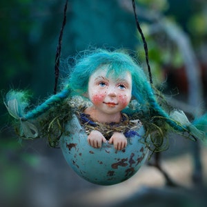 May include: A charming doll with turquoise hair and rosy cheeks is nestled in a hanging, weathered blue eggshell. The doll's hands are visible, and the eggshell is suspended by a dark string. The doll is surrounded by foliage, creating a fairy-tale aesthetic.