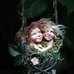 Könnte beinhalten: Eine skurrile Skulptur von zwei Figuren, die in einem moosbedeckten Nest sitzen und an einem Zweig hängen. Die Figuren haben rosige Wangen und fließendes Haar, eines rot und eines blond. Das Nest ist mit kleinen weißen Blumen und grünem Laub geschmückt.