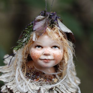 May include: A fairy ornament with a smiling face, blonde hair, and freckles. The fairy's head is surrounded by a skirt of dried petals and adorned with leaves and purple accents. The ornament is suspended by a thin wire.