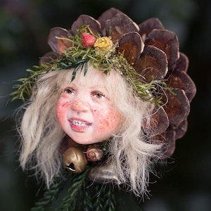 Könnte beinhalten: Ein skurriles Weihnachtsschmuckstück mit einem lächelnden Gesicht mit blonden Haaren, Sommersprossen und einem Tannenzapfen-Kopfschmuck. Der Schmuck ist mit Grün, kleinen Glocken und Beeren verziert und schafft eine festliche und einzigartige Weihnachtsdekoration.