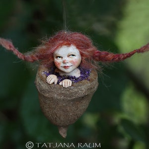 May include: A handmade fairy figure with red hair and a smiling face, emerging from a brown acorn-shaped pod. The fairy is wearing a purple top. The background is a blurred green.