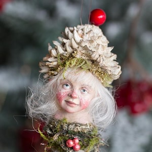 May include: A handmade Christmas ornament featuring a whimsical figure with a cone-shaped head adorned with pinecone scales and a small red berry. The figure has a painted face, white hair, and is surrounded by greenery. A red berry is at the top and bottom.