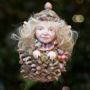 May include: A detailed fairy ornament with a smiling face, blonde hair, and a detailed costume. The fairy is set within a pinecone base, holding small, colourful flowers. The background is a blurred green.