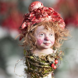 May include: A handcrafted decorative figure with a whimsical design. The figure features a doll-like face with rosy cheeks, a red floral headdress, and a textured white body. The figure is adorned with natural elements like moss and berries.