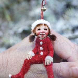 May include: A charming Christmas elf ornament, dressed in a red jumpsuit with white detailing and buttons. The elf has a white hat, brown pigtails, and a smiling face. A small bell hangs from the top. A unique festive decoration.