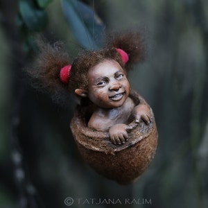 May include: A handcrafted figurine of a smiling child with brown skin, curly brown hair, and red hair ties, emerging from a brown, textured shell. The figurine is suspended against a blurred green background.