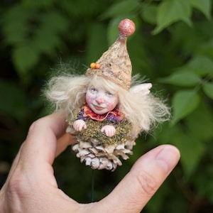 May include: A handmade fairy ornament with a whimsical design. The fairy has a painted face, blonde hair, and a pointed hat. It is nestled in a pinecone base, with a mossy texture. The fairy's outfit includes a red and purple collar.