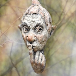 Op de afbeelding: Een gedetailleerde sculptuur van een kabouterachtig gezicht met een vinger op de lippen, wat stilte suggereert. Het gezicht is getextureerd met tinten grijs, bruin en beige, met zichtbare rimpels en gedetailleerde kenmerken. De sculptuur staat tegen een vage natuurlijke achtergrond.