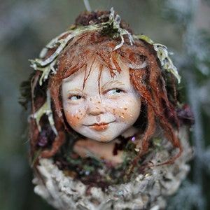 May include: A detailed, handcrafted fairy ornament. The fairy has red hair, freckles, and a gentle smile, with a leafy crown. The ornament is set in a textured, earthy base. A unique piece for festive decoration, with intricate details.