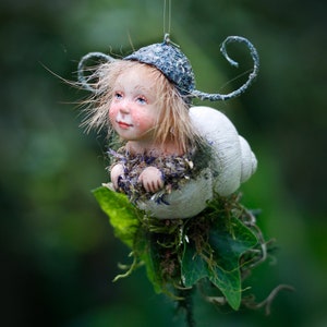 Puede incluir: Una caprichosa figura de hada con cuerpo de concha de caracol, que lleva un sombrero azul con detalles rizados. El hada tiene el pelo rubio y mejillas rosadas, rodeada de vegetación. La figura está suspendida por un hilo fino.