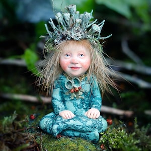 K&ouml;nnte beinhalten: Eine skurrile Figur einer sitzenden Fee in einem tealfarbenen Kleid und einer Krone aus nat&uuml;rlichen Elementen. Die Fee hat blondes Haar, rosige Wangen und blaue Augen. Die Figur befindet sich in einer nat&uuml;rlichen, moosigen Umgebung.
