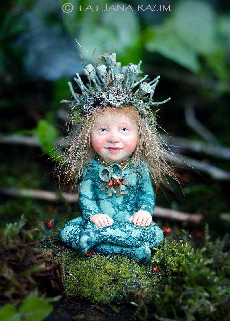 K&ouml;nnte beinhalten: Eine skurrile Figur einer sitzenden Fee in einem tealfarbenen Kleid und einer Krone aus nat&uuml;rlichen Elementen. Die Fee hat blondes Haar, rosige Wangen und blaue Augen. Die Figur befindet sich in einer nat&uuml;rlichen, moosigen Umgebung.