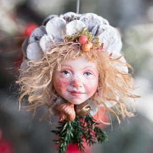 May include: A whimsical Christmas ornament featuring a doll face with blonde hair, blue eyes, and rosy cheeks. The ornament has a white, petal-like headdress, a sprig of greenery, and small bells. The artist's name, TATJANA RAUM, is visible at the bottom.