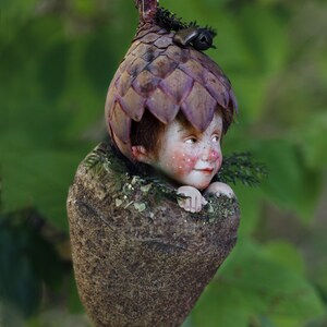 Può includere: Una stravagante statuetta di fata fatta a mano. La fata ha una testa dettagliata a forma di ghianda con un piccolo viso e un corpo marrone strutturato. La fata è sospesa da sottili fili su uno sfondo verde sfocato.