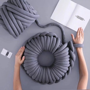 May include: A gray chunky yarn is being used to create a large, round, gray knitted pillow. The yarn is being wrapped around the pillow to create a circular shape. The pillow is being made on a gray surface. The text "HOW TO MAKE AN OCHO WITH CHUNKY YARN" is visible on a white piece of paper.