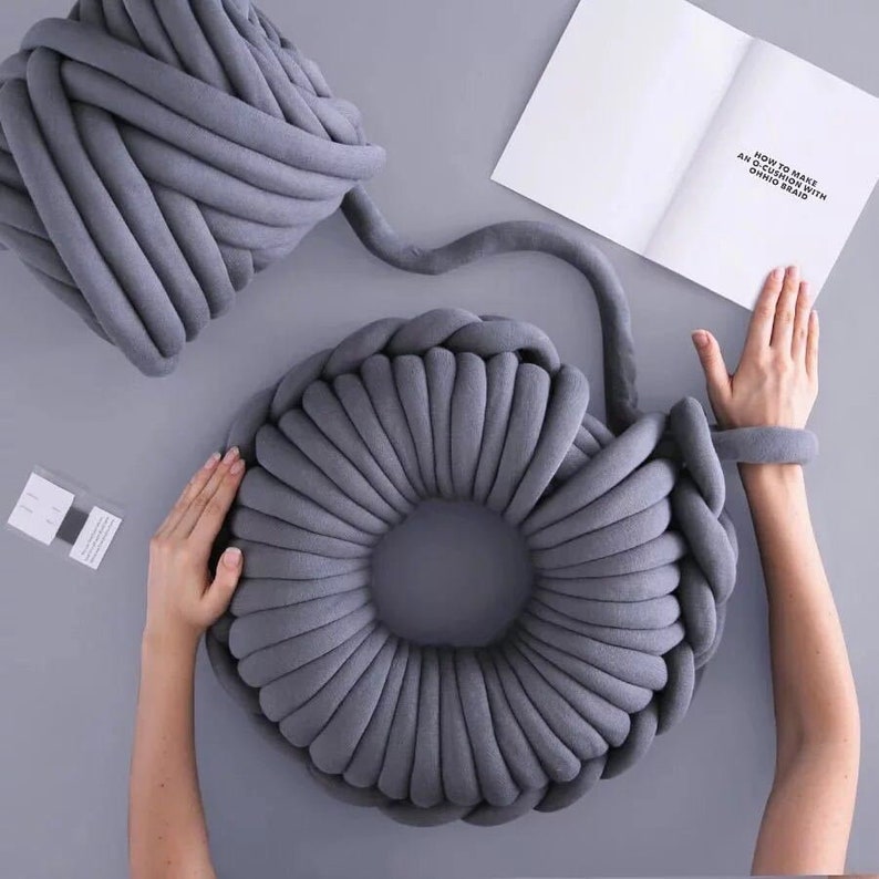 May include: A gray chunky yarn is being used to create a large, round, gray knitted pillow. The yarn is being wrapped around the pillow to create a circular shape. The pillow is being made on a gray surface. The text "HOW TO MAKE AN OCHO WITH CHUNKY YARN" is visible on a white piece of paper.