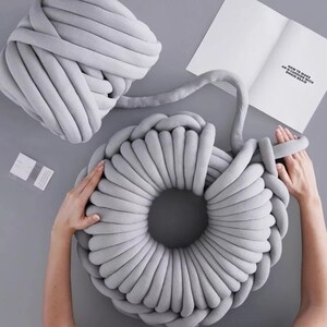 May include: A gray chunky yarn is being used to create a large, round, gray knitted pillow. The yarn is being wrapped around itself to create the pillow. The pillow is being made on a gray surface. The text "HOW TO MAKE A CUSHION WITH CHUNKY YARN" is visible on a white piece of paper.