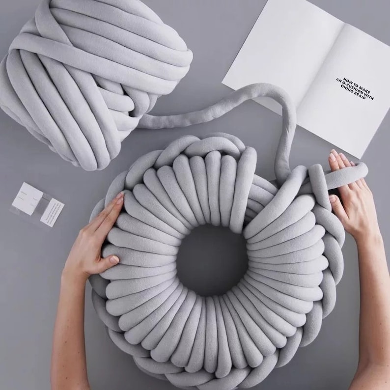 May include: A gray chunky yarn is being used to create a large, round, gray knitted pillow. The yarn is being wrapped around itself to create the pillow. The pillow is being made on a gray surface. The text "HOW TO MAKE A CUSHION WITH CHUNKY YARN" is visible on a white piece of paper.