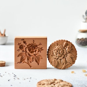 May include: A wooden cookie cutter and a cookie with a detailed rose design. The cookie cutter is made of light brown wood and has a square shape. The cookie is round and has a brown color.