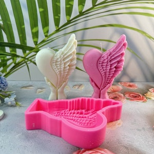 May include: Two heart-shaped candles with angel wings, one cream and one pink, are displayed. A pink silicone mould in the shape of a heart with wings is in the foreground. The candles are on a light grey surface.