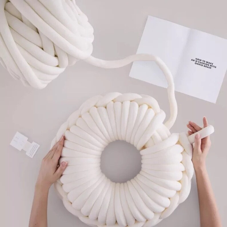 May include: A white, chunky yarn is being used to create a large, round, white cushion. The yarn is being braided into a circular shape. The instructions for making the cushion are visible in the background. "How to make an O-cushion with chunky braid"