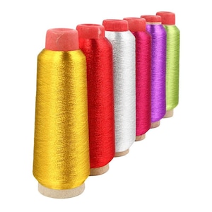 Op de afbeelding: Zes spoelen met metallic garen in verschillende kleuren, waaronder goud, rood, zilver, roze, paars en groen. De spoelen zijn in een rij gerangschikt, met de gouden spoel vooraan.