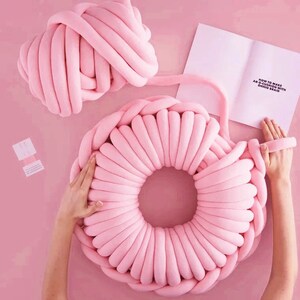 May include: A pink, large, round, knitted cushion is being made by hand. The cushion is made from a thick, chunky yarn. The yarn is being braided around the cushion to create a circular shape. The instructions for making the cushion are visible in the background. The instructions are titled "How to Make an XL Cushion with Chunky Braid".